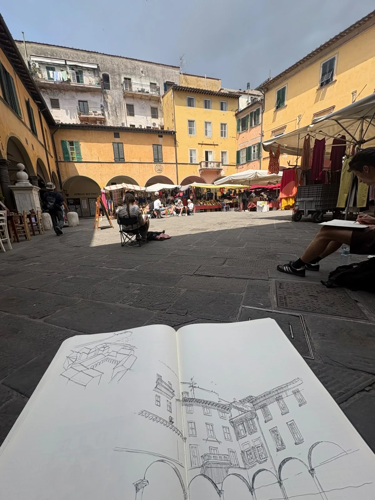 An open sketchbook resting on stone paving, showing a pen drawing of an arcaded Italian market square, with the real square and its market stalls beyond.