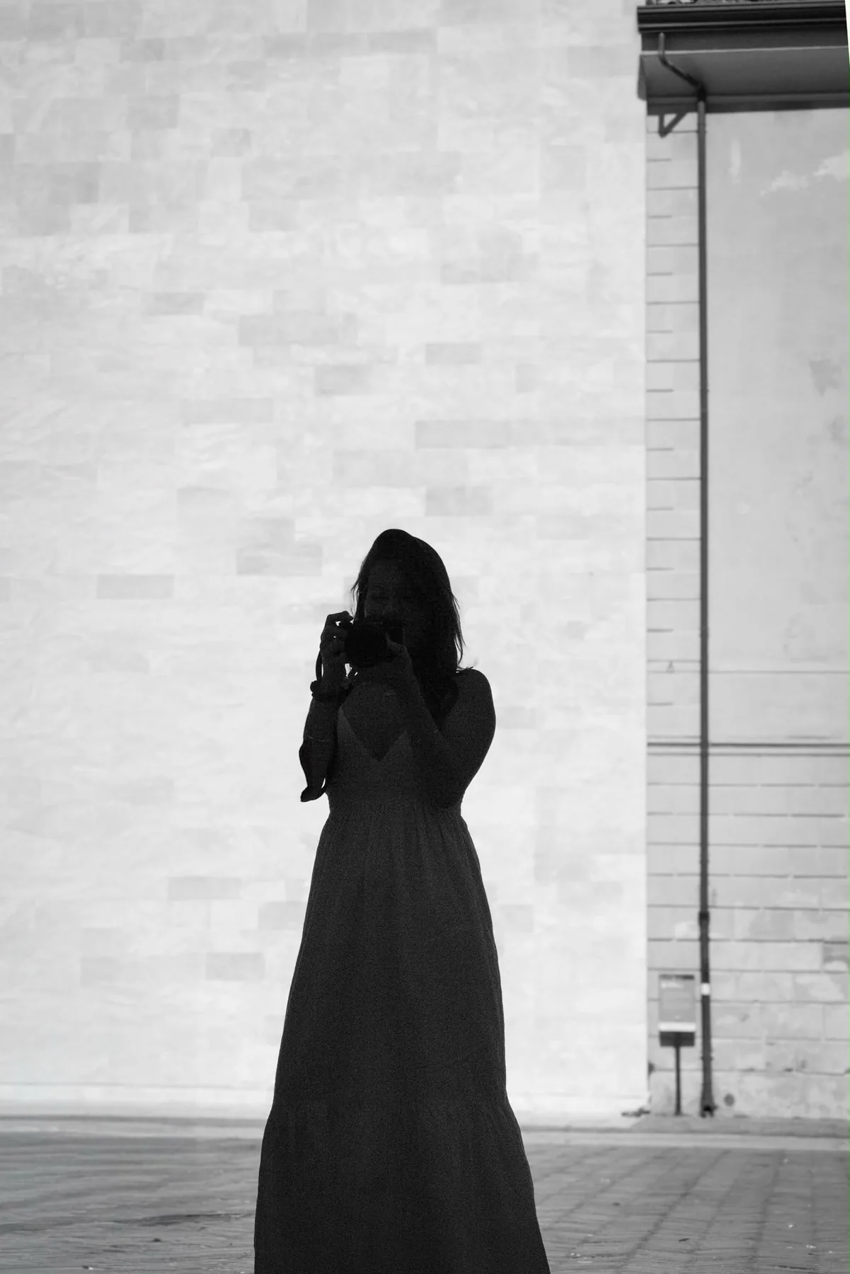 Sofie Saymé photographed in silhouette against a pale stone wall, holding a camera to her face.